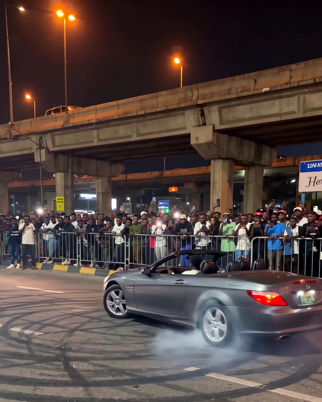 A night street takeover under a Lagos flyover, a crowd behind metal barricades, tyre smoke on wet asphalt